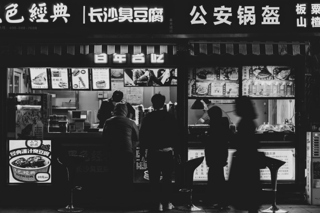 Silhouettes of people at a street food stall at night with vibrant signage.